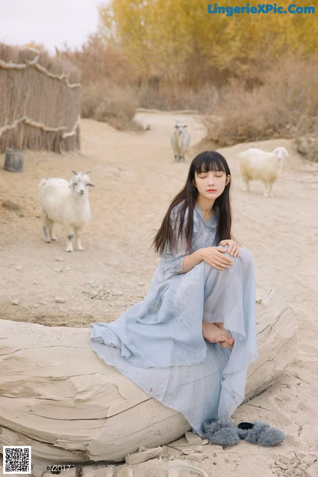 A woman sitting on a log in front of a herd of goats.